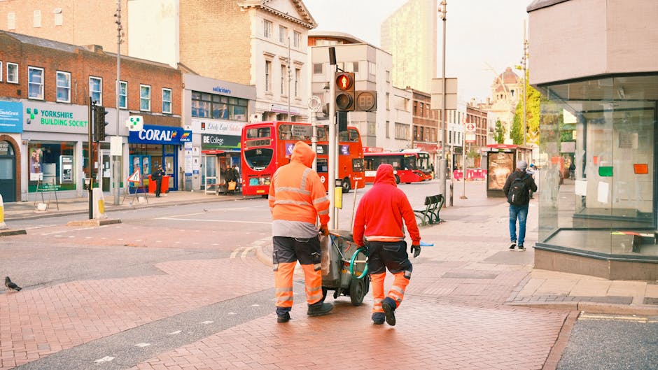 Two workers dressed in high-visibility orange and red uniforms are performing surface cleaning on a city street in Watford. They are pushing equipment across the red-brick sidewalk near a traffic ligh